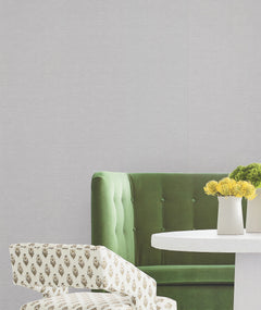 A green velvet sofa and a patterned chair are arranged beside a white round table with two small potted yellow flowers, set against walls adorned with York Wallcoverings’ Hempfield Weave Loomed Wallpaper.