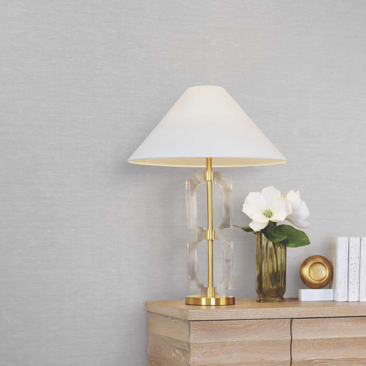 A gold and glass table lamp with a white shade sits on a wooden dresser next to a vase with white flowers, a gold decorative object, and several white books, set against walls covered in York Wallcoverings’ Hempfield Weave Loomed Wallpaper.