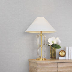 A gold and glass table lamp with a white shade sits on a wooden dresser next to a vase with white flowers, a gold decorative object, and several white books, set against walls covered in York Wallcoverings’ Hempfield Weave Loomed Wallpaper.