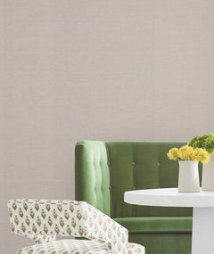 A green tufted sofa, a round white table with two white vases of yellow flowers, and a patterned upholstered chair are arranged against a wall covered in Hempfield Weave Loomed Wallpaper by York Wallcoverings.