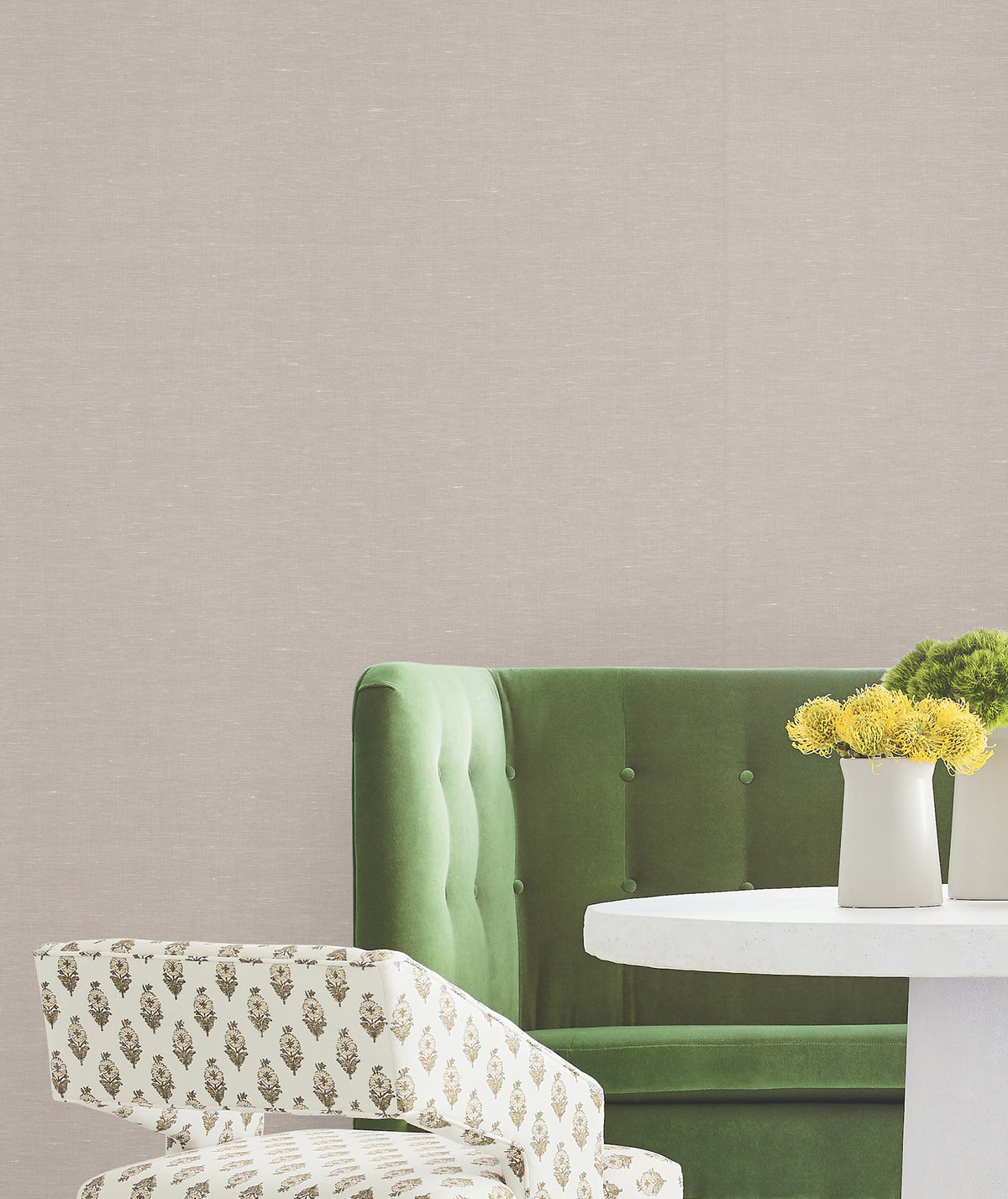Green velvet sofa, round white table with two vases of yellow flowers, and a patterned chair, set against a plain beige wall adorned with Hempfield Weave Loomed Wallpaper by York Wallcoverings.