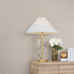 A modern lamp with a white shade sits on a wooden table next to a vase with white flowers, a small gold dish, and a few stacked white books, all set against walls covered in Hempfield Weave Loomed Wallpaper by York Wallcoverings.