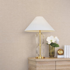 A modern lamp with a white shade sits on a wooden table next to a gold vase with white flowers, a gold candle holder, and three stacked white books against a wall covered in York Wallcoverings’ Hempfield Weave Loomed Wallpaper.