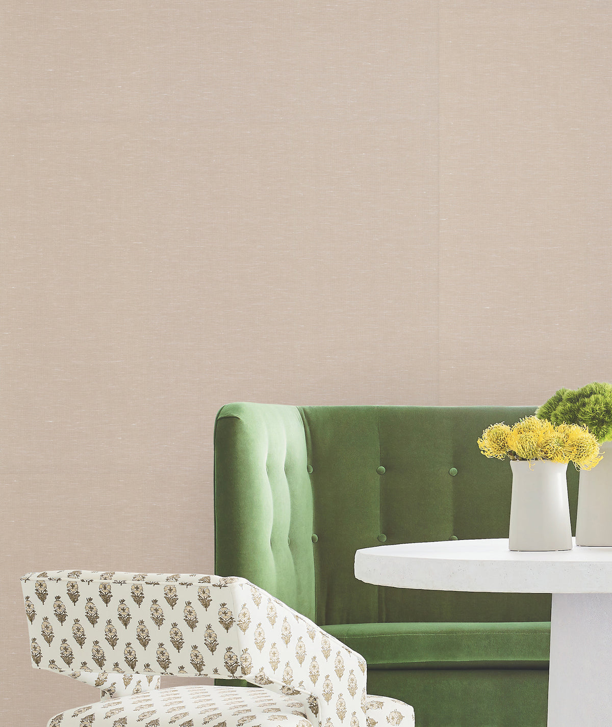 A green tufted sofa, a white chair with a floral pattern, and a round white table with yellow flowers in a beige vase are set against walls covered in York Wallcoverings’ Hempfield Weave Loomed Wallpaper.