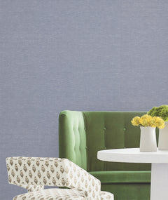 A green sofa, a patterned chair, a round white table, and a vase with yellow flowers are set against a light blue York Wallcoverings Hempfield Weave Loomed Wallpaper.
