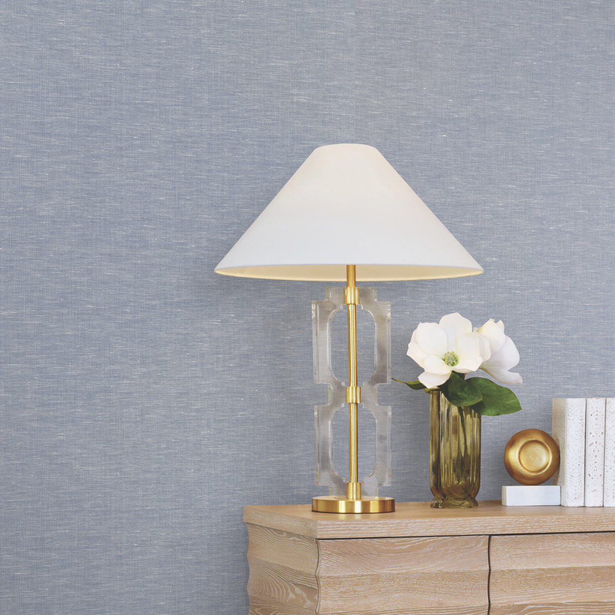 A gold and glass table lamp with a white shade sits on a wooden dresser beside white flowers in a gold vase, a gold-rimmed dish, and stacked white books, all set against a backdrop of York Wallcoverings’ Hempfield Weave Loomed Wallpaper.
