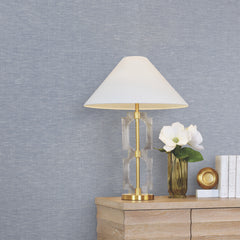 A gold and glass table lamp with a white shade sits on a wooden dresser beside white flowers in a gold vase, a gold-rimmed dish, and stacked white books, all set against a backdrop of York Wallcoverings’ Hempfield Weave Loomed Wallpaper.