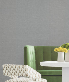 A green velvet sofa and a patterned white chair are arranged beside a round white table topped with yellow flowers in a beige vase, all set against a gray wall adorned with Hempfield Weave Loomed Wallpaper from York Wallcoverings.