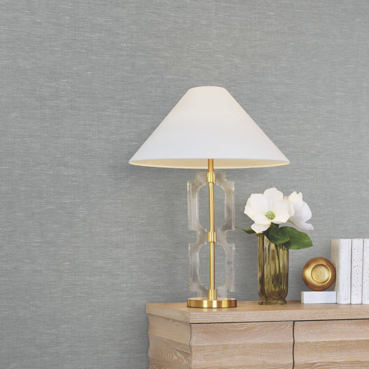 A gold and clear table lamp with a white shade sits on a wooden dresser next to a white flower in a vase, a gold decorative item, and a few stacked books against a wall adorned with Hempfield Weave Loomed Wallpaper by York Wallcoverings.
