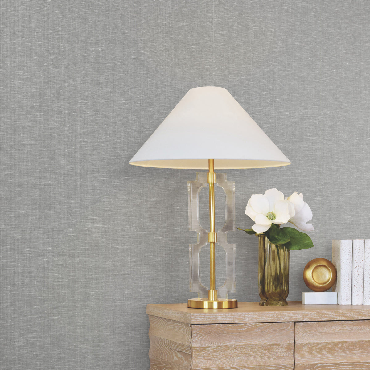 A modern table lamp with a white shade and gold base sits on a wooden dresser next to a vase with white flowers, a gold decorative object, and stacked white books, all set against a wall covered in York Wallcoverings’ Hempfield Weave Loomed Wallpaper.