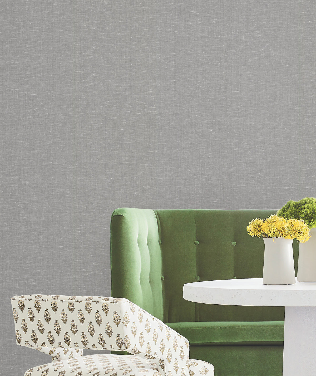 A green velvet sofa, a white table with two white vases holding yellow flowers, and a patterned chair are set against a backdrop of York Wallcoverings’ Hempfield Weave Loomed Wallpaper.