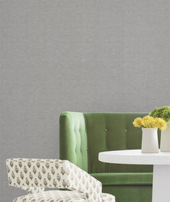 A green velvet sofa, a white table with two white vases holding yellow flowers, and a patterned chair are set against a backdrop of York Wallcoverings’ Hempfield Weave Loomed Wallpaper.