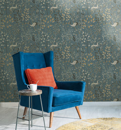 A blue armchair with an orange cushion sits beside a side table with a white mug, set against Aspen Wallpaper by York Wallcoverings, featuring stylized deer, birds, and botanical motifs.
