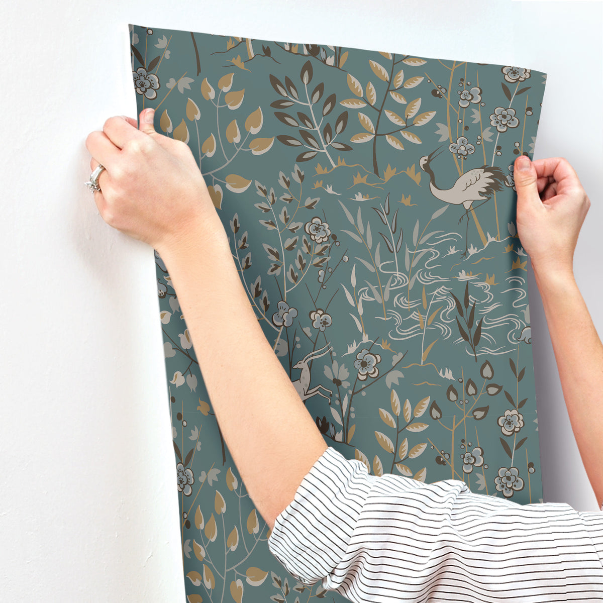 Hands applying a sheet of teal Aspen Wallpaper by York Wallcoverings, featuring a pattern of stylized cranes, flowers, and foliage, to a wall.
