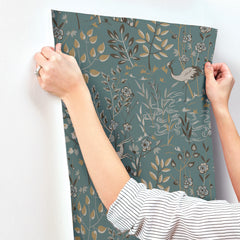 Hands applying a sheet of teal Aspen Wallpaper by York Wallcoverings, featuring a pattern of stylized cranes, flowers, and foliage, to a wall.