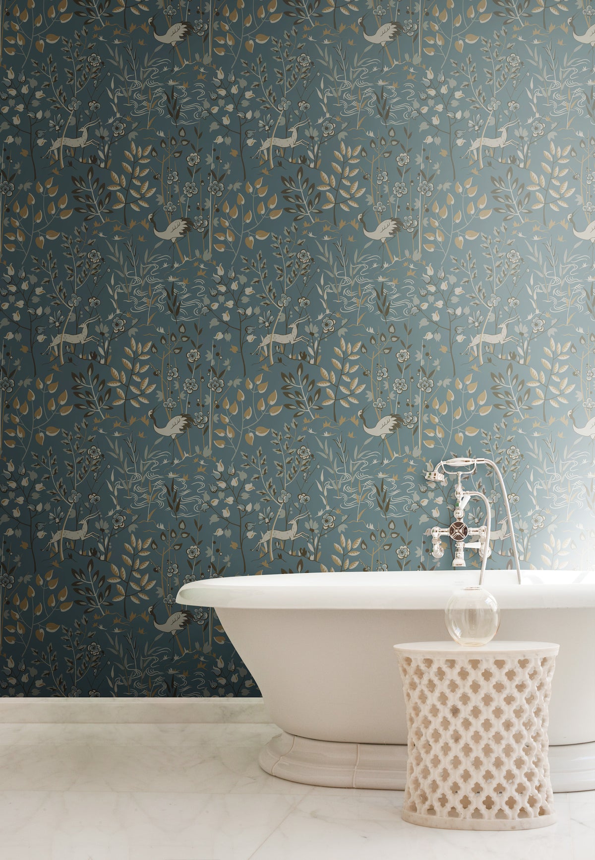 A white clawfoot bathtub and a patterned side table stand in front of York Wallcoverings Aspen Wallpaper, which features repeating illustrations of deer, birds, and botanical motifs.