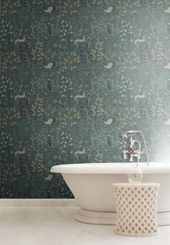 A white clawfoot bathtub and a patterned side table stand in front of York Wallcoverings Aspen Wallpaper, which features repeating illustrations of deer, birds, and botanical motifs.