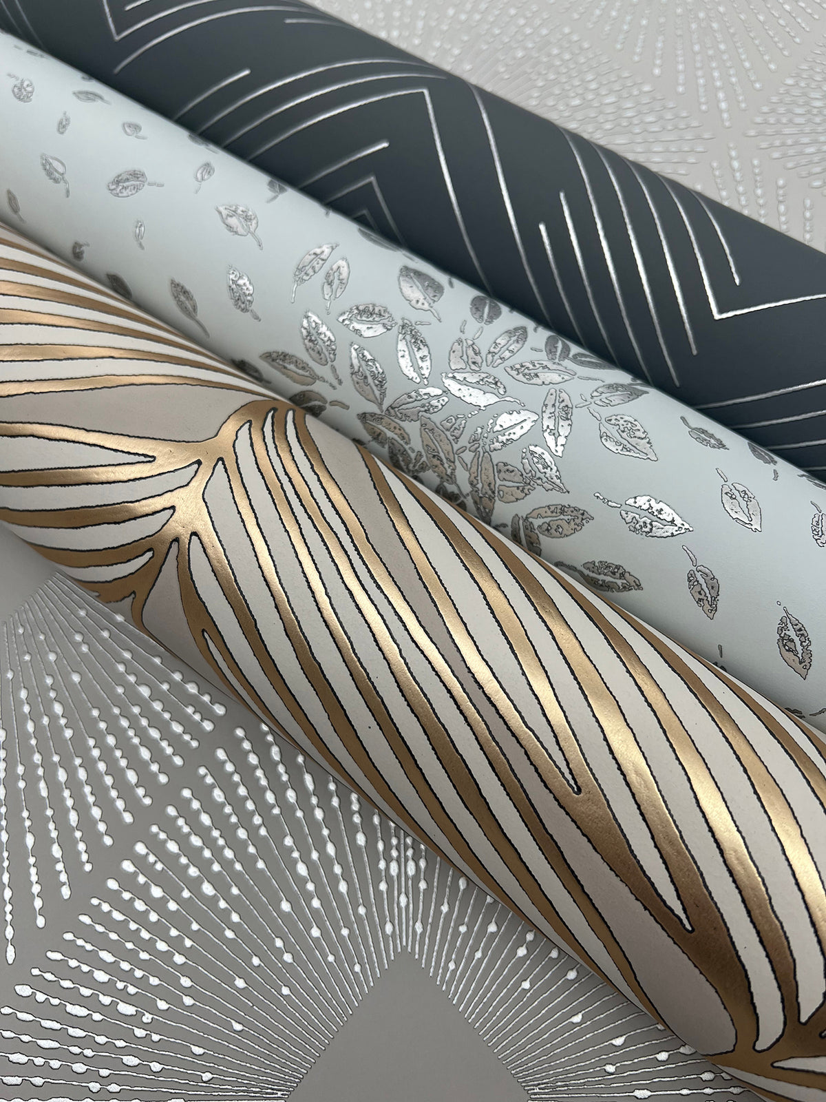 Three rolls of York Wallcoverings Petite Leaves Wallpaper featuring metallic designs—dark grey with silver lines, light grey with silver leaves, and white with gold and silver abstract lines—rest on patterned paper adorned with silver dots and lines.