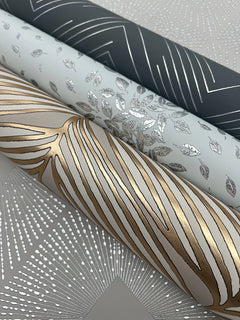Three rolls of York Wallcoverings Petite Leaves Wallpaper featuring metallic designs—dark grey with silver lines, light grey with silver leaves, and white with gold and silver abstract lines—rest on patterned paper adorned with silver dots and lines.