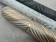 Three rolls of decorative wallpaper by York Wallcoverings, including the Petite Leaves Wallpaper, featuring metallic silver and gold patterns: one with radiating lines, one with delicate leaves, and one with bold gold stripes on white.