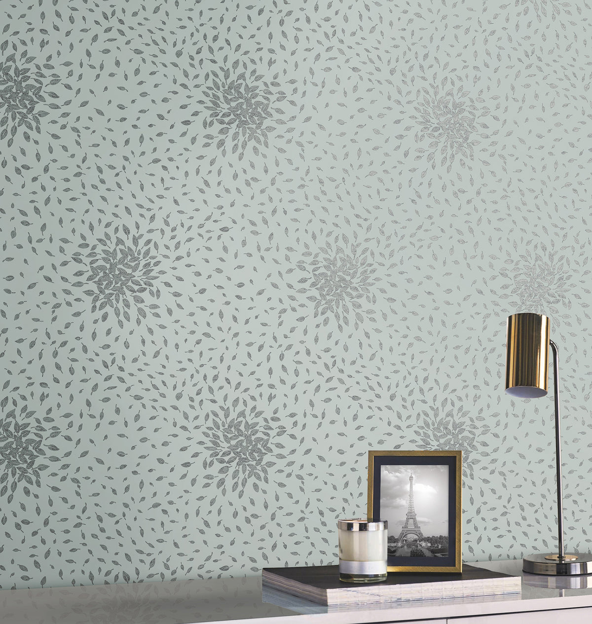 A desk with a gold lamp, a candle, and a framed black-and-white photo of the Eiffel Tower sits in front of a light blue wall adorned with York Wallcoverings’ Petite Leaves Wallpaper, featuring a repeating pattern of gray, leaf-like shapes forming starbursts.