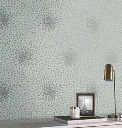A desk with a gold lamp, a candle, and a framed black-and-white photo of the Eiffel Tower sits in front of a light blue wall adorned with York Wallcoverings’ Petite Leaves Wallpaper, featuring a repeating pattern of gray, leaf-like shapes forming starbursts.