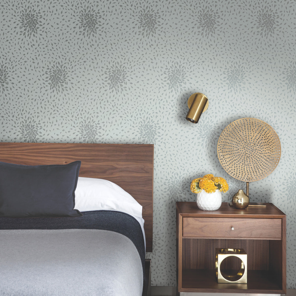 Modern bedroom with a wooden headboard, gray bedding, and a nightstand holding a vase of yellow flowers, gold decor, and a woven accent. The walls feature Petite Leaves Wallpaper by York Wallcoverings, complemented by a gold wall sconce in the background.