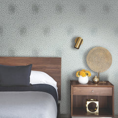 Modern bedroom with a wooden headboard, gray bedding, and a nightstand holding a vase of yellow flowers, gold decor, and a woven accent. The walls feature Petite Leaves Wallpaper by York Wallcoverings, complemented by a gold wall sconce in the background.