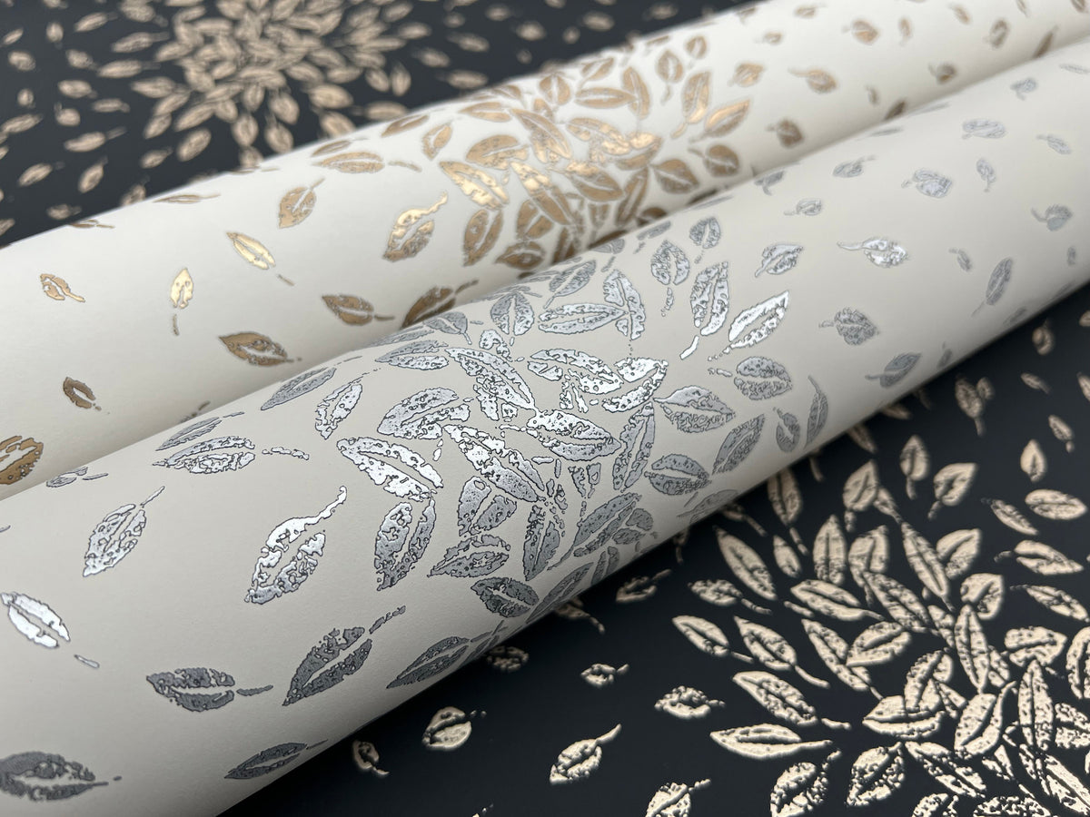 Three rolls of Petite Leaves Wallpaper by York Wallcoverings, featuring metallic silver and gold leaf patterns on cream and black backgrounds, displayed at an angle atop a surface with a coordinating leafy design.