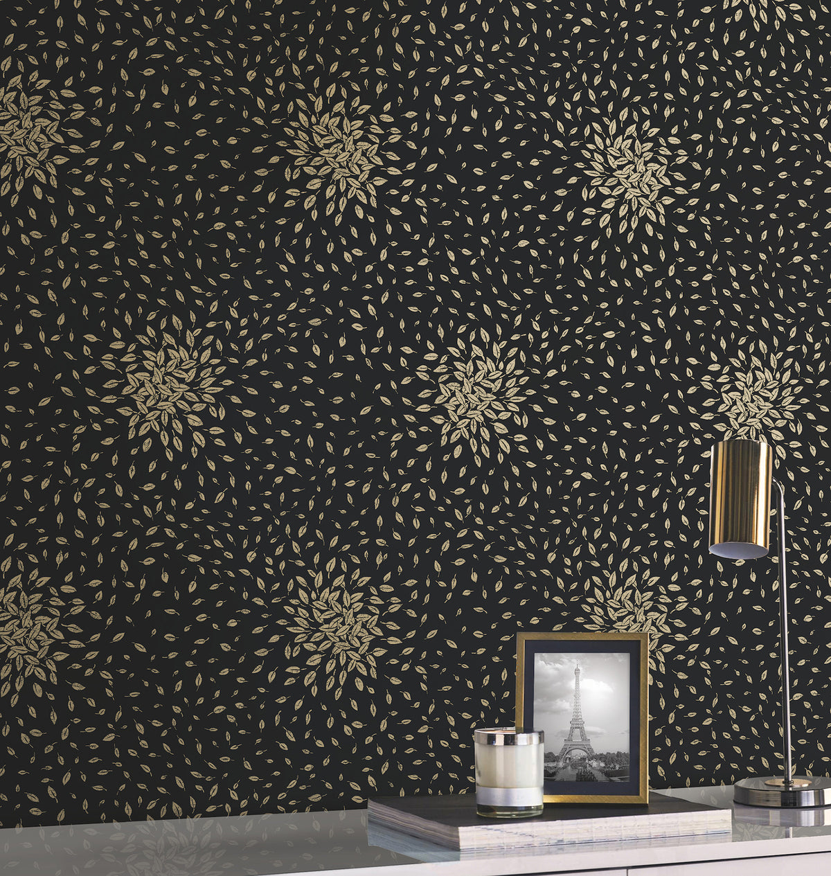 A wall covered in Petite Leaves Wallpaper by York Wallcoverings, featuring a gold leaf pattern on black; a white tabletop with a gold lamp, a framed photo of the Eiffel Tower, and a small silver container.