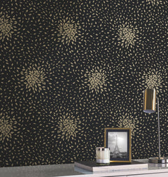 A wall covered in Petite Leaves Wallpaper by York Wallcoverings, featuring a gold leaf pattern on black; a white tabletop with a gold lamp, a framed photo of the Eiffel Tower, and a small silver container.