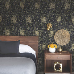 A modern bedroom featuring York Wallcoverings’ Petite Leaves Wallpaper as a dark floral-patterned accent wall, complemented by a wooden headboard, gray bedding, gold wall lamp, nightstand, a vase of yellow flowers, and decorative gold accents.