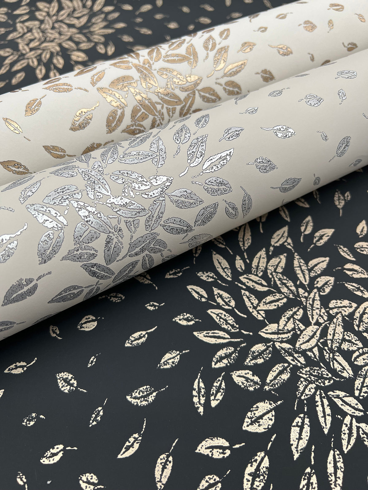 Three rolls of Petite Leaves Wallpaper by York Wallcoverings—in gold, silver, and bronze metallic leaf patterns—are displayed on a black surface adorned with complementary leaf motifs, creating an elegant, nature-inspired aesthetic.