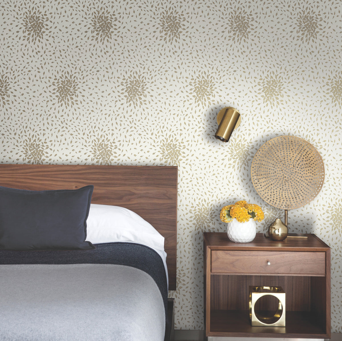 Modern bedroom with a wooden bed, gray bedding, and a nightstand. On the nightstand are a decorative woven lamp, a vase with yellow flowers, and a gold object. The wall features the Petite Leaves Wallpaper by York Wallcoverings in a gold floral pattern.