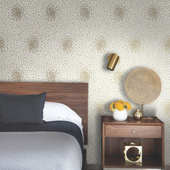 Modern bedroom with a wooden bed, gray bedding, and a nightstand. On the nightstand are a decorative woven lamp, a vase with yellow flowers, and a gold object. The wall features the Petite Leaves Wallpaper by York Wallcoverings in a gold floral pattern.