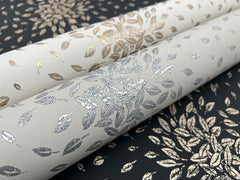 Three rolls of York Wallcoverings Petite Leaves Wallpaper, each featuring a metallic leaf pattern in gold or silver on white and black backgrounds. The designs are arranged in a circular, radiating motif.