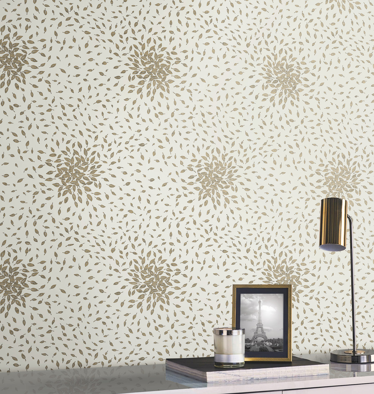 A modern desk is styled with a gold lamp, a framed black-and-white photo of the Eiffel Tower, and stacked books. Behind, York Wallcoverings’ Petite Leaves Wallpaper adds elegance with its gold floral pattern on a light background.