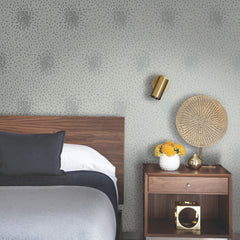 A modern bedroom featuring a wooden bed and nightstand, gray bedding, a gold wall lamp, and a vase of yellow flowers, set against York Wallcoverings’ Petite Leaves Wallpaper.