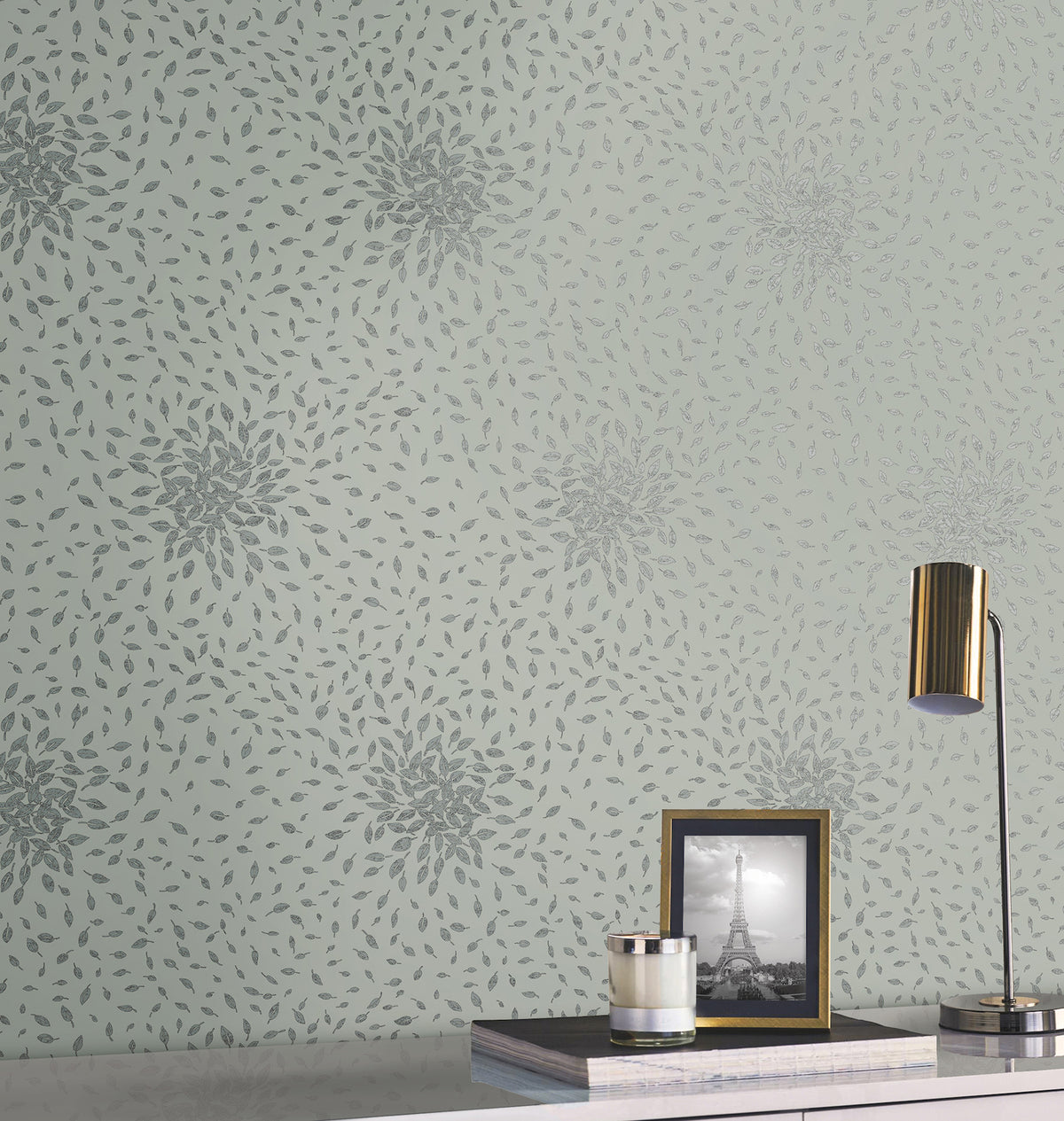 A wall adorned with York Wallcoverings’ Petite Leaves Wallpaper in pale green showcases a subtle floral leaf pattern. Resting on a white shelf are a gold lamp, a candle, and a framed black-and-white photo of the Eiffel Tower.