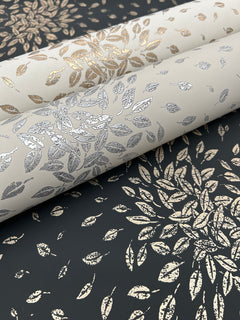 Three rolls of Petite Leaves Wallpaper by York Wallcoverings, featuring intricate metallic leaf patterns in gold and silver tones, are displayed on a dark background with coordinating leaf designs. The wallpaper’s detailed motifs create a luxurious and elegant look.