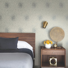 A modern bedroom with a wooden headboard, gray bedding, Petite Leaves Wallpaper by York Wallcoverings, a nightstand holding a vase of yellow flowers, a golden lamp, and a decorative round wicker piece.