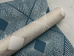 A close-up of Harlowe Wallpaper by York Wallcoverings, showcasing its metallic geometric patterns with radiating lines and diamond shapes in blue, white, and gold tones. A roll of the wallpaper rests on top of the patterned background.