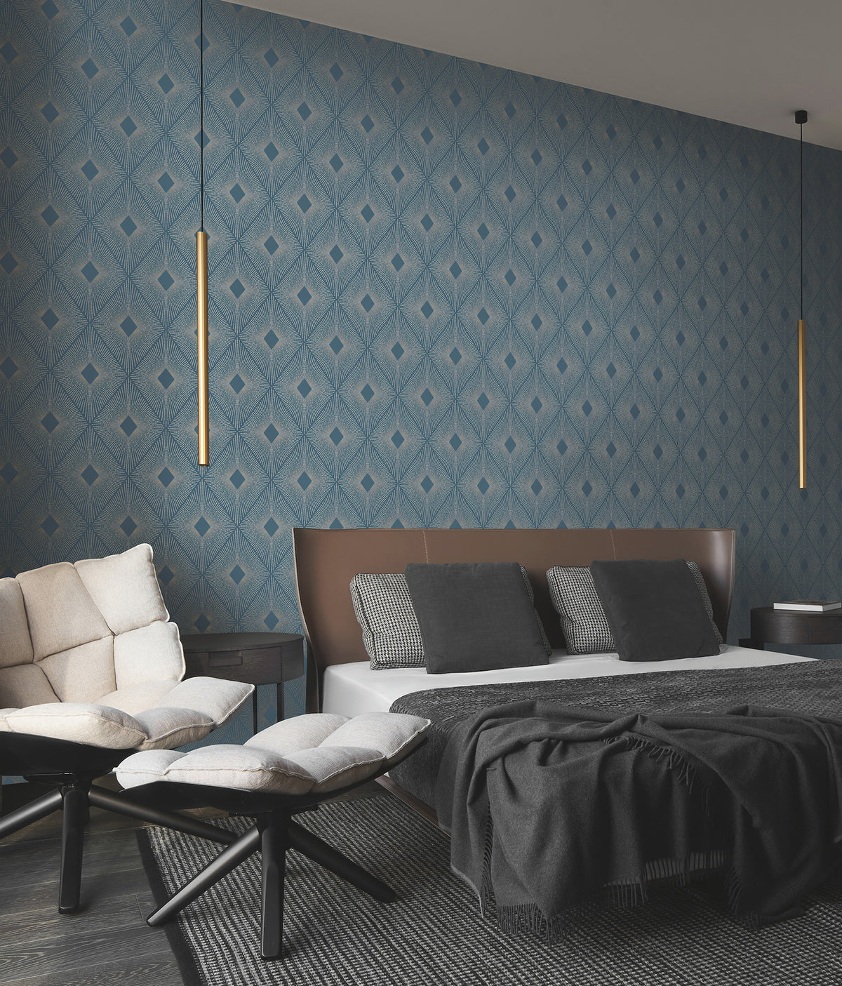 Modern bedroom featuring Harlowe Wallpaper by York Wallcoverings in a geometric blue design, paired with a brown headboard, gray bedding, black and white pillows, cushioned chairs, and two slim hanging pendant lights flanking the bed. Dark wood floors complete the look.