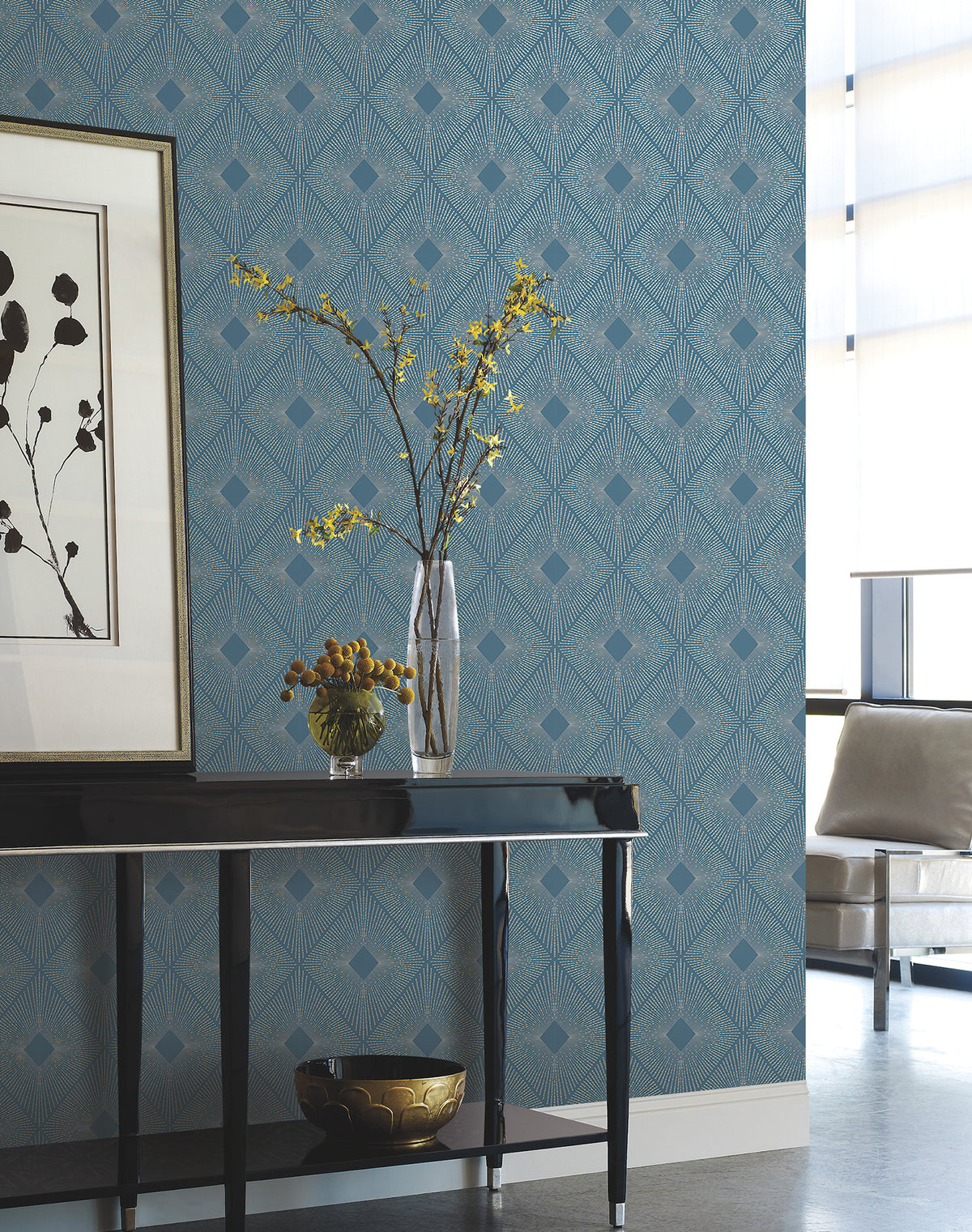 A modern room featuring Harlowe Wallpaper by York Wallcoverings with a black console table, a vase of yellow flowers, a bowl of fruit, wall art, and a beige chair by a window adorned with sheer curtains.