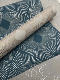 A close-up of rolled and unrolled Harlowe Wallpaper by York Wallcoverings, showcasing a geometric starburst pattern in blue, gold, and silver hues, with metallic dotted lines radiating from central squares.