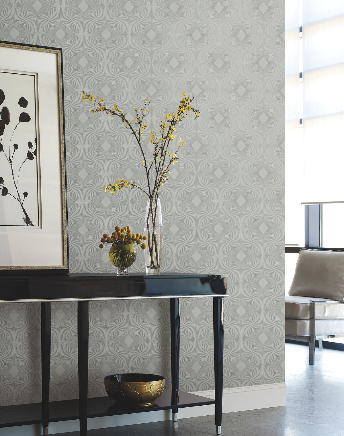 A modern living room featuring a black console table, glass vase with yellow flowers, decorative bowl, framed artwork, and Harlowe Wallpaper by York Wallcoverings in a geometric gray pattern. Sunlight filters through sheer window coverings.