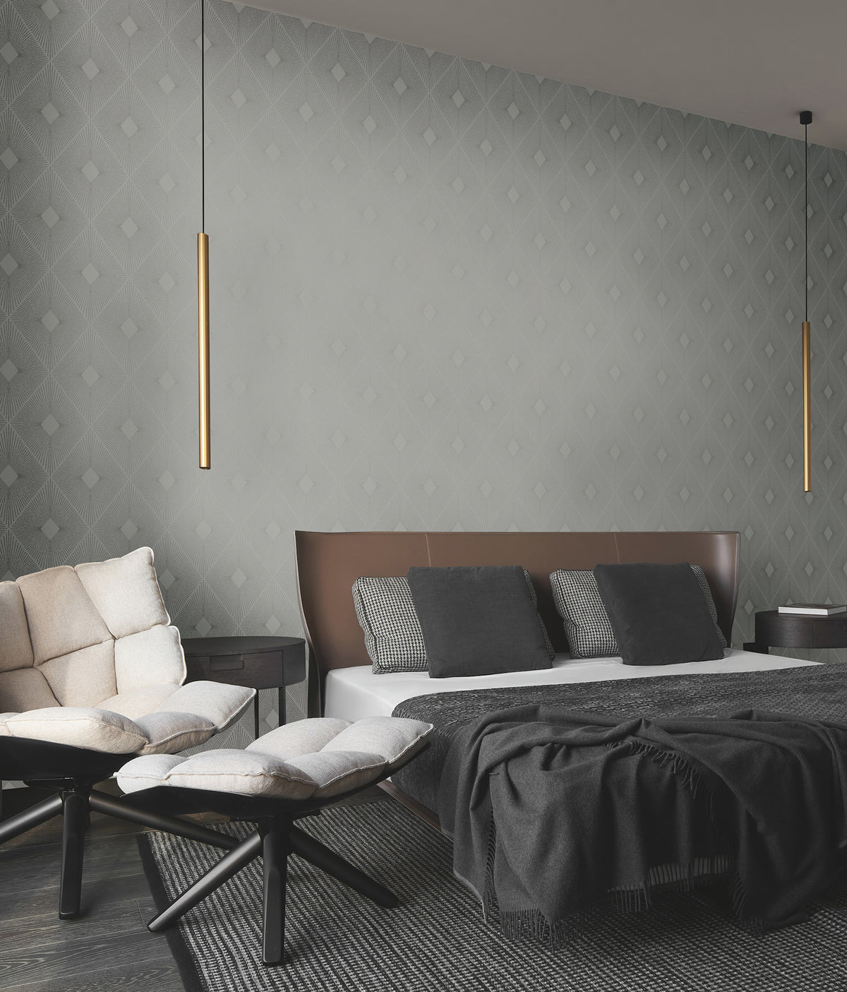Modern bedroom featuring a large bed with gray bedding, a cushioned bench, two round ottomans, and Harlowe Wallpaper by York Wallcoverings as the patterned backdrop, complemented by two long gold pendant lights hanging from the ceiling.
