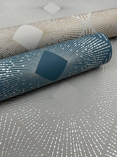 Three rolls of Harlowe Wallpaper by York Wallcoverings are shown close up, featuring a geometric sunburst pattern in metallic gold and silver on gray and teal backgrounds. The designs showcase radiating lines emerging from a central diamond shape.