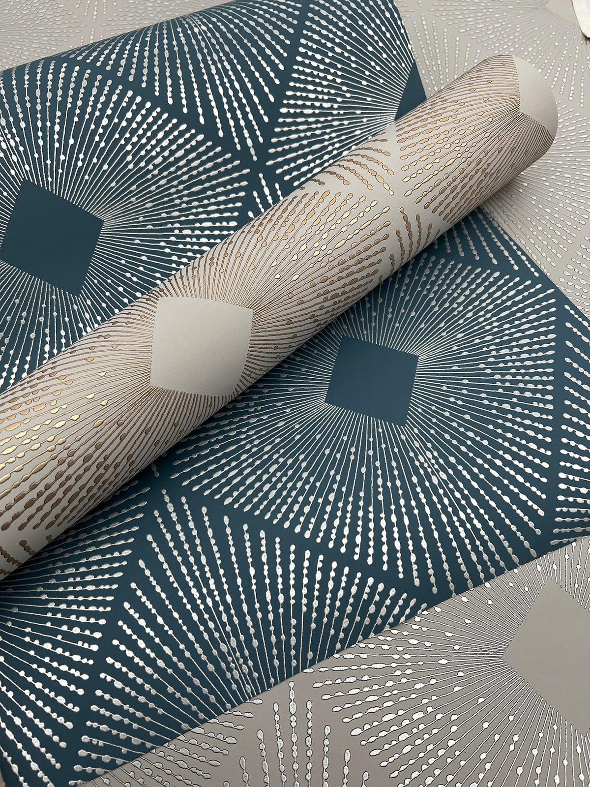 The Harlowe Wallpaper by York Wallcoverings features a geometric teal and silver starburst pattern with metallic accents, shown partially unrolled on a coordinating background.