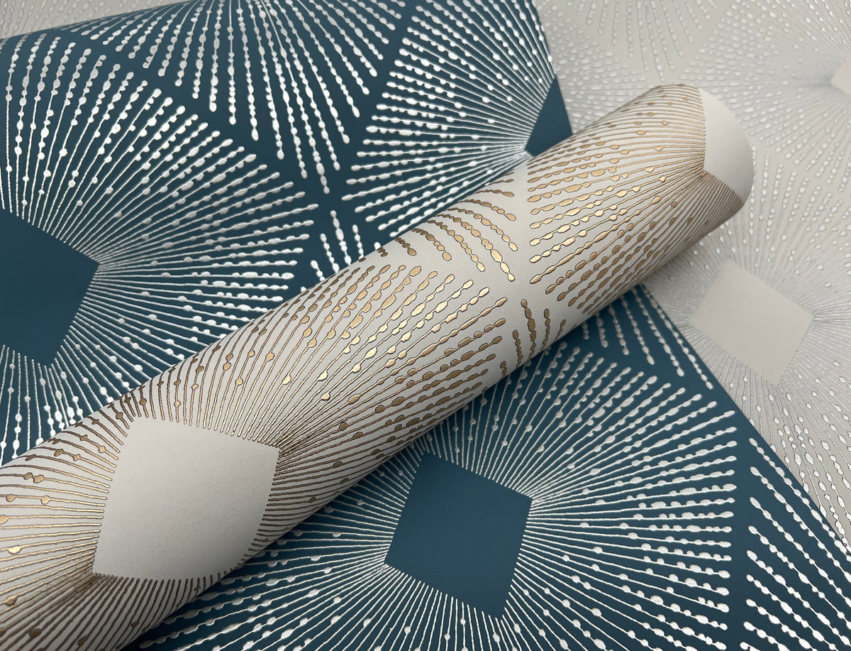 A close-up of the Harlowe Wallpaper by York Wallcoverings, showcasing decorative geometric patterns with metallic gold and silver lines radiating from square shapes on a dark teal and light background. A partially unrolled roll of the wallpaper is visible.